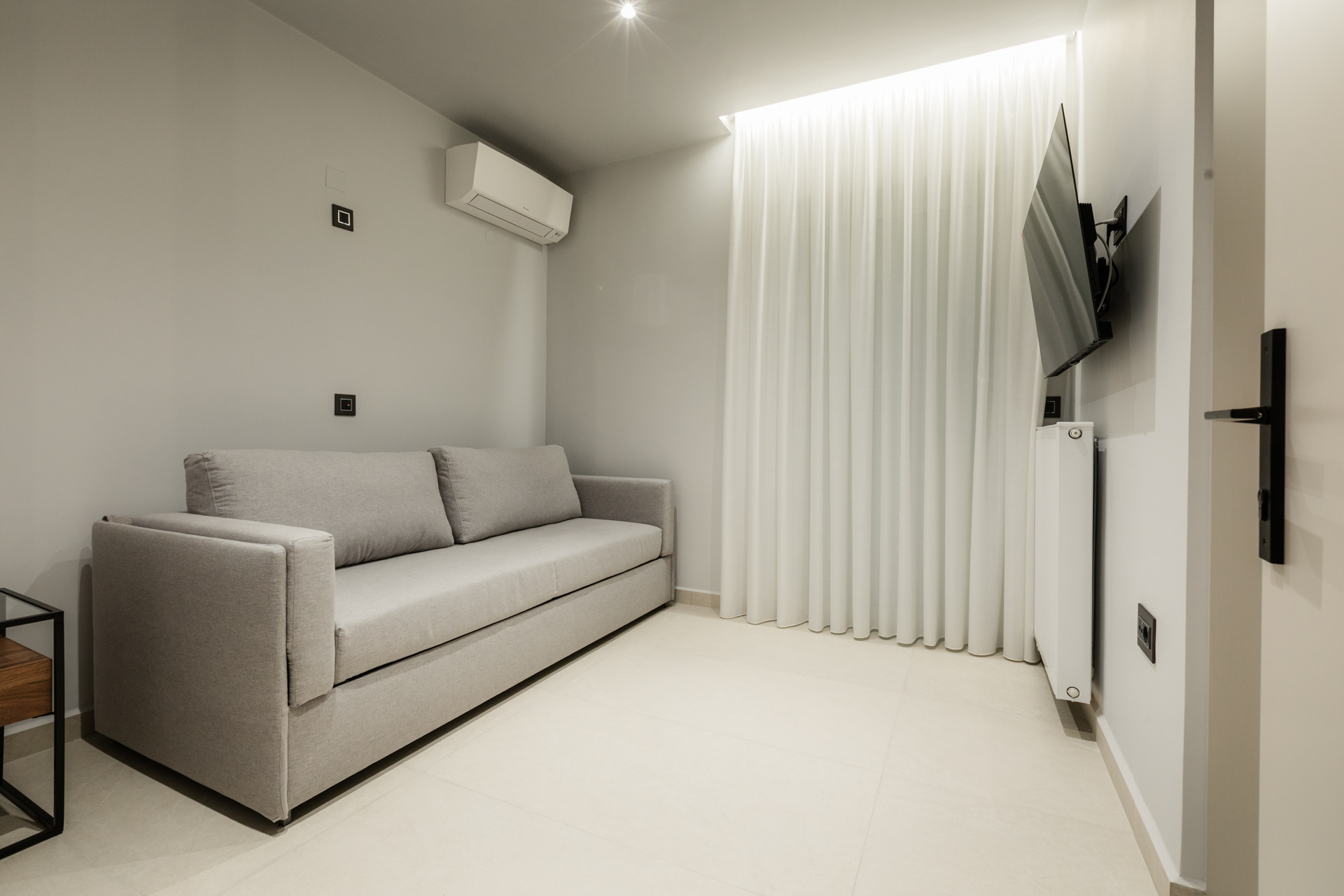 Minimal ξενώνας με γκρι καναπέ-κρεβάτι, λευκές κουρτίνες με κρυφό φωτισμό και τηλεόραση τοίχου."